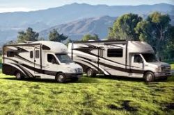 Picture of 2 different RVs parked on green grass with trees and mountains in the background.